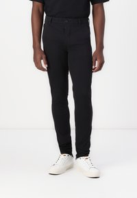 Black fitted pants made of a smooth, stretchy fabric with a button closure and belt loops, paired with white sneakers and a black shirt.