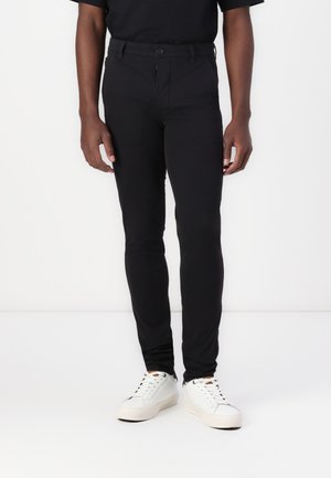 Black fitted pants made of a smooth, stretchy fabric with a button closure and belt loops, paired with white sneakers and a black shirt.