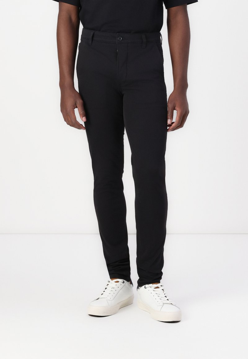 Black fitted pants made of a smooth, stretchy fabric with a button closure and belt loops, paired with white sneakers and a black shirt.