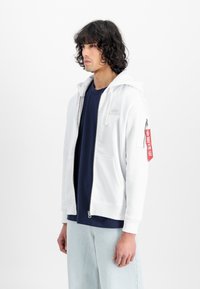 Sudadera blanca con cremallera y capucha, bolsillos frontales y un detalle de etiqueta roja en la manga. Llevada sobre una camiseta azul marino y pantalones de mezclilla claros.