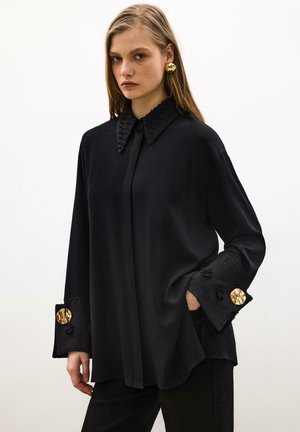 Mlada žena u širokoj crnoj bluzi s teksturiranim ovratnikom i manžetama, ukrašenim velikim zlatnim okruglim gumbima, stoji uz jednobojnu pozadinu.