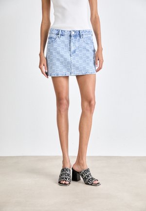 Falda vaquera azul claro con un sutil patrón de logos por todo el tejido, con cierre de botones y bolsillos estándar. Combinada con sandalias tipo slide negras con logo.