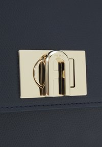 Borsa a tracolla in pelle texturizzata blu navy con chiusura dotata di gancio in metallo dorato, caratterizzata da marchio in rilievo e dettaglio a loop decorativo.