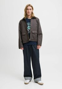 Brun lynlåsjakke med spids krave, to forlommer, båret over en mørk skjorte og parret med brede mørke jeans og hvide sneakers.