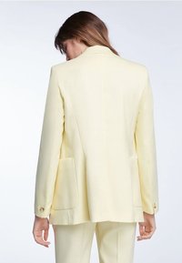 Vaaleankeltainen räätälöity bleiseri, jossa on yksirivinen malli, kaksi etutaskua ja napituskoristelu, joka tarjoaa sileän tekstuurin ja rakenteellisen istuvuuden.