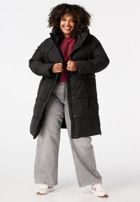 Zwarte lange pufferjas met capuchon, voorzien van een gewatteerd ontwerp, knopen aan de voorkant en zijzakken, gedragen over een bordeauxrood topje en grijze jeans.