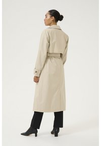 Bež trenč kaput s pojasom u struku, dugim rukavima i skraćenim dizajnom straga. Uparen s crnim hlačama i crnim cipelama s oštrim vrhom.