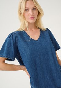 Džinsu tunika ar puff piedurknēm, V veida kaklu un brīvu piegriezumu. Iezīmējas ar vidēji zilu krāsu ar smalku tekstūru un redzamām šuvēm.