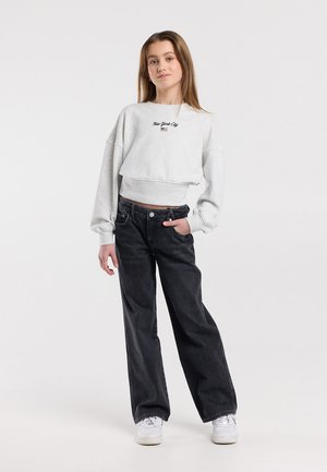 Tienermeisje staat met hand in zak, draagt een lichtgrijze "New York City"-hoodie, zwarte jeans met brede pijpen en witte sneakers.