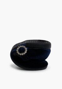 Zapatilla de terciopelo azul marino con forma redondeada, suela texturada negra y un acento decorativo en forma de círculo plateado en la correa.