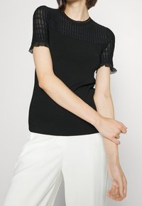 Černý vypasovaný top s krátkými rukávy z průsvitné ribbed síťky, kulatým výstřihem a kraťounkým lemováním. Spárován s bílými kalhotami s širokými nohavicemi.