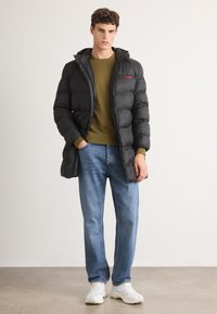 Chaqueta acolchada negra con capucha, con un diseño acolchado y logo rojo. Combinada con un suéter verde oliva, jeans azul claro y zapatillas blancas.