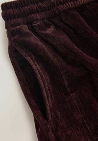 Burgunderfarbene Samthose mit weicher Textur, die einen elastischen Bund und seitliche Taschen aufweisen, aus einem schrägen Blickwinkel präsentiert.