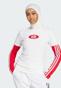 Bela rebrasta majica z rdečimi dolgimi rokavi, ki ima tri bele stripe in rdeči vezeni logotip na prsih, v kombinaciji z belimi hlačami.