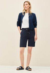 Blouson de pilote bleu marine avec des poignets élastiques, t-shirt blanc à col rond, shorts bleu marine avec taille à cordon de serrage, et sandales foncées avec un design de sangle.