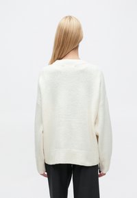 Kremfarget, strikket genser med avslappet passform, ribbet kant og vide ermer, som viser en myk tekstur. Brukt med mørke, skreddersydde bukser.