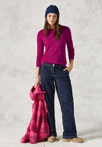 Top a maniche lunghe fucsia, jeans a gamba larga blu scuro e un berretto in maglia blu navy. Tiene un giubbotto a scacchi rosa. Texture morbide e colori vivaci.