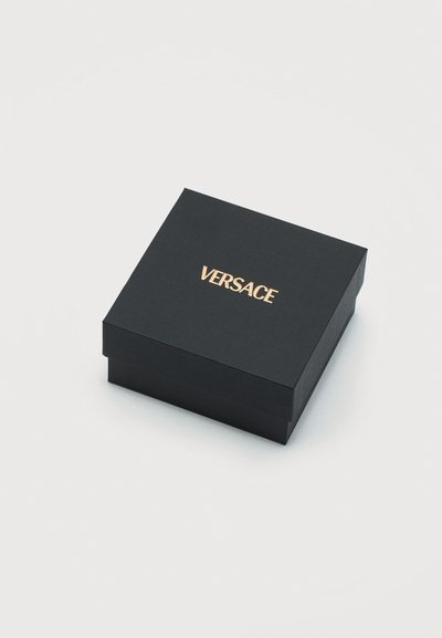 Μαύρο τετράγωνο κουτί δώρου με υφή στην επιφάνεια και χρυσό λογότυπο "VERSACE" στο κέντρο του καπακιού, σε απλό ανοιχτό φόντο.