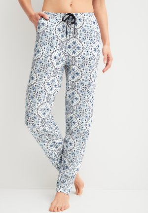 Persoon die witte en blauwe broek met bloemmotief en trekkoord draagt, staat op blote voeten met één hand in de zak tegen een effen achtergrond.