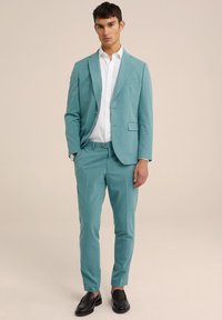 Traje turquesa con un blazer de un solo botón y pantalones ajustados, combinado con una camisa blanca y mocasines negros. Textura de tela suave.
