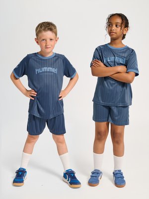 Dwoje dzieci w dopasowanych niebieskich strojach sportowych stoi obok siebie, jedno z rękami na biodrach, drugie z założonymi rękami, na białym tle.