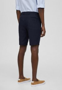 Pantaloni corti in lino blu navy con una tasca posteriore dotata di chiusura a bottone, abbinati a scarpe slip-on color beige e una camicia a righe chiare.