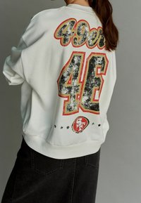 Felpa bianca con design grafico rosso e nero, con "49ers" e "46" sul retro. Texture morbida e vestibilità rilassata. Gonna di jeans nera sotto.