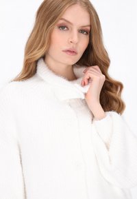 White knitted cardigan with a soft, textured finish. Features a wide collar and button closure. Loose fit with long sleeves.