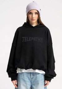 Czarna, oversize bluza z kapturem z napisem "TELEPATHY" nadrukowanym w pikselowej czcionce. Posiada sznurek w kapturze oraz szerokie, ściągacze przy rękawach. Nosi się ją na białej koszulce.