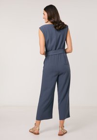 Navyblauer Overall mit weitem Bein, tailliertem Bund und Kurzarm. Glatte Textur und minimalistisches Design, im Rückenansicht.