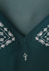 Tessuto teal con foglie ricamate in argento lungo il collo, con un dettaglio a forma di diamante al centro. Morbido al tatto, design a forma di V.
