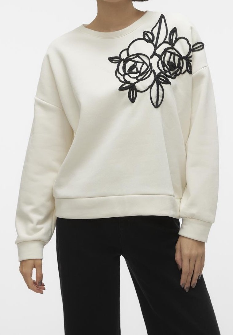 Femme portant un sweat-shirt blanc avec une broderie florale noire à l'épaule, associé à un pantalon noir.
