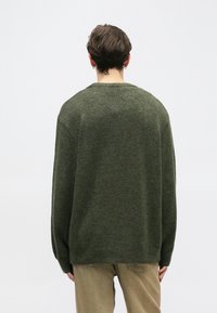 Pulover supradimensionat verde, dintr-un material moale de tricot, cu gât rotund și tiv canelat. Purtați-l cu pantaloni de culoare maro deschis.
