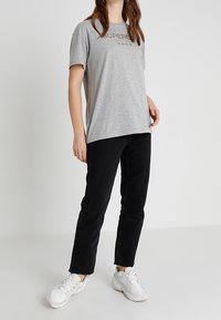 Camiseta de algodón gris con mangas cortas que presenta el texto "SUPER" en dorado, combinada con pantalones vaqueros negros de pierna recta y zapatillas blancas.