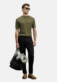 Olijfgroene ribgebreide sweater met korte mouwen, zwarte maatspijkerbroek, bruine suède schoenen, donkere zonnebril, en twee tassen in contrasterende kleuren.