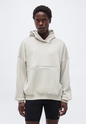 Modelo masculino con sudadera oversized de color blanco roto con bolsillo frontal con cremallera y pantalones cortos de ciclismo negros, de pie contra un fondo liso.