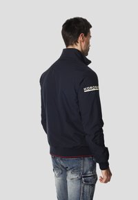 Námořnicky modrá bunda s vysokým límcem, elastickými manžetami a spodním lemem; na levém rukávu je bílý nápis "KOROSHI". Navržena s hladkou texturou.