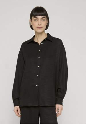 Kvinde med kort mørkt hår iført en løs sort skjorte med knapper og matchende sorte bukser, stående mod en ensfarvet lys baggrund.