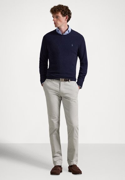 Marinblå crewneck-tröja, beige chinos, bruna loafers; tyget ser mjukt och texturerat ut. Tröjan har en liten logga och en skräddarsydd passform.