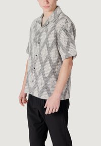 Camicia a maniche corte con bottoni di colore grigio chiaro e un motivo geometrico nero. Realizzata in un tessuto intrecciato testurizzato, presenta un colletto a camicia e una vestibilità rilassata.