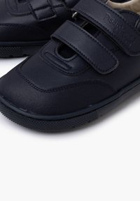 Zapatos infantiles de cuero negro con correas de velcro, superficie texturizada, punta redondeada y suela de goma con un agarre en patrón.