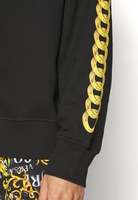 Chemise noire à manches longues avec un imprimé chaîne dorée courant sur la manche, portée avec un pantalon noir à motifs jaunes et blancs.