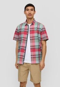 Camicia a maniche corte a quadretti nei toni del rosa, verde e nero, con colletto button-down e vestibilità dritta, indossata sopra una t-shirt bianca e pantaloni corti beige.