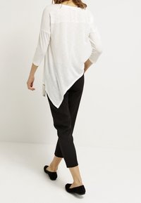 Weißes asymmetrisches Tunika-Oberteil mit dreiviertel Ärmeln, strukturiertem Stoff, fließendem Design, kombiniert mit schwarzen verkürzten Hosen und schwarzen Ballerinas.
