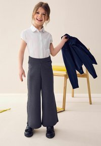 Pantalons larges gris, chemise blanche à manches courtes, tenant une veste bleu marine dans une pièce lumineuse avec une chaise en bois et des objets jaunes.