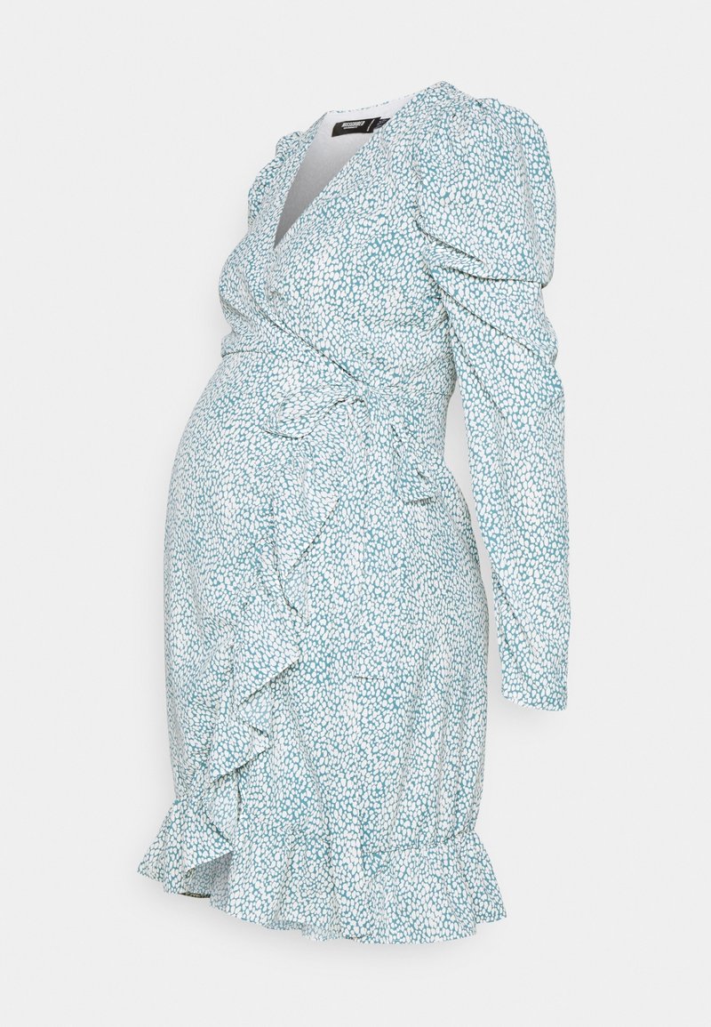 Sukienka ciążowa w jasno-niebieskim kolorze z białym wzorem w kropki. Posiada fason kopertowy, długie rękawy oraz falbaniasty dół, dodający tekstury.