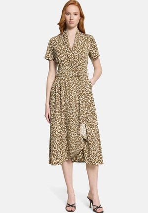 Femme aux cheveux roux portant une robe portefeuille mi-longue à imprimé léopard marron et beige, et des sandales noires à talons, debout avec une main dans la poche.