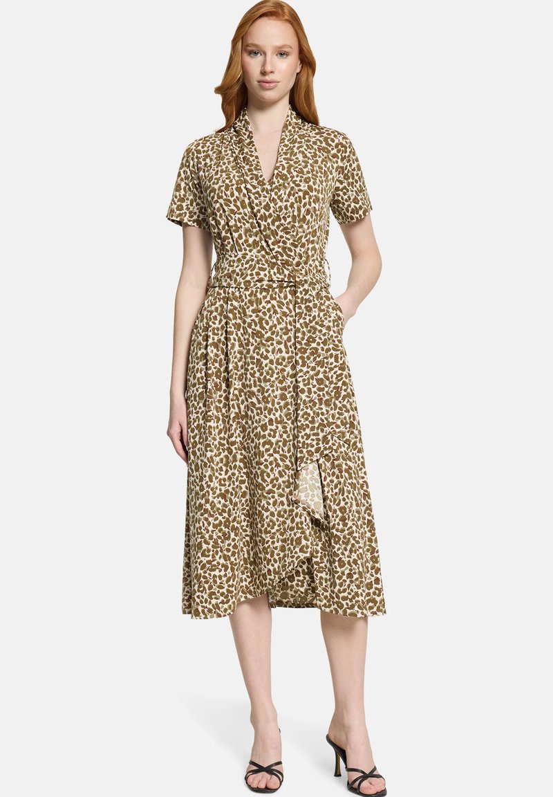 Femme aux cheveux roux portant une robe portefeuille mi-longue à imprimé léopard marron et beige, et des sandales noires à talons, debout avec une main dans la poche.