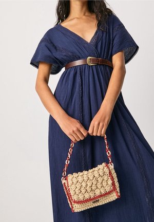 Mujer vestida con un vestido azul marino y un cinturón marrón, sosteniendo un bolso tejido beige con ribete rojo y asa trenzada.