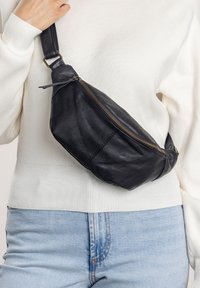 Schwarze Lederbauchtasche mit runder Form, goldener Reißverschluss und verstellbarem Tragegurt, getragen über einem weißen Pullover und hellblauen Jeans.